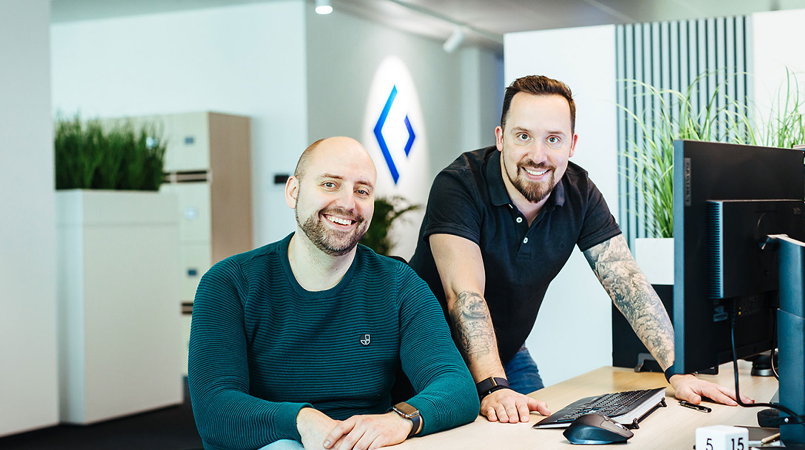 Olaf Malinowski und Mario Techel sitzen zusammen am Schreibtisch und lächeln. 