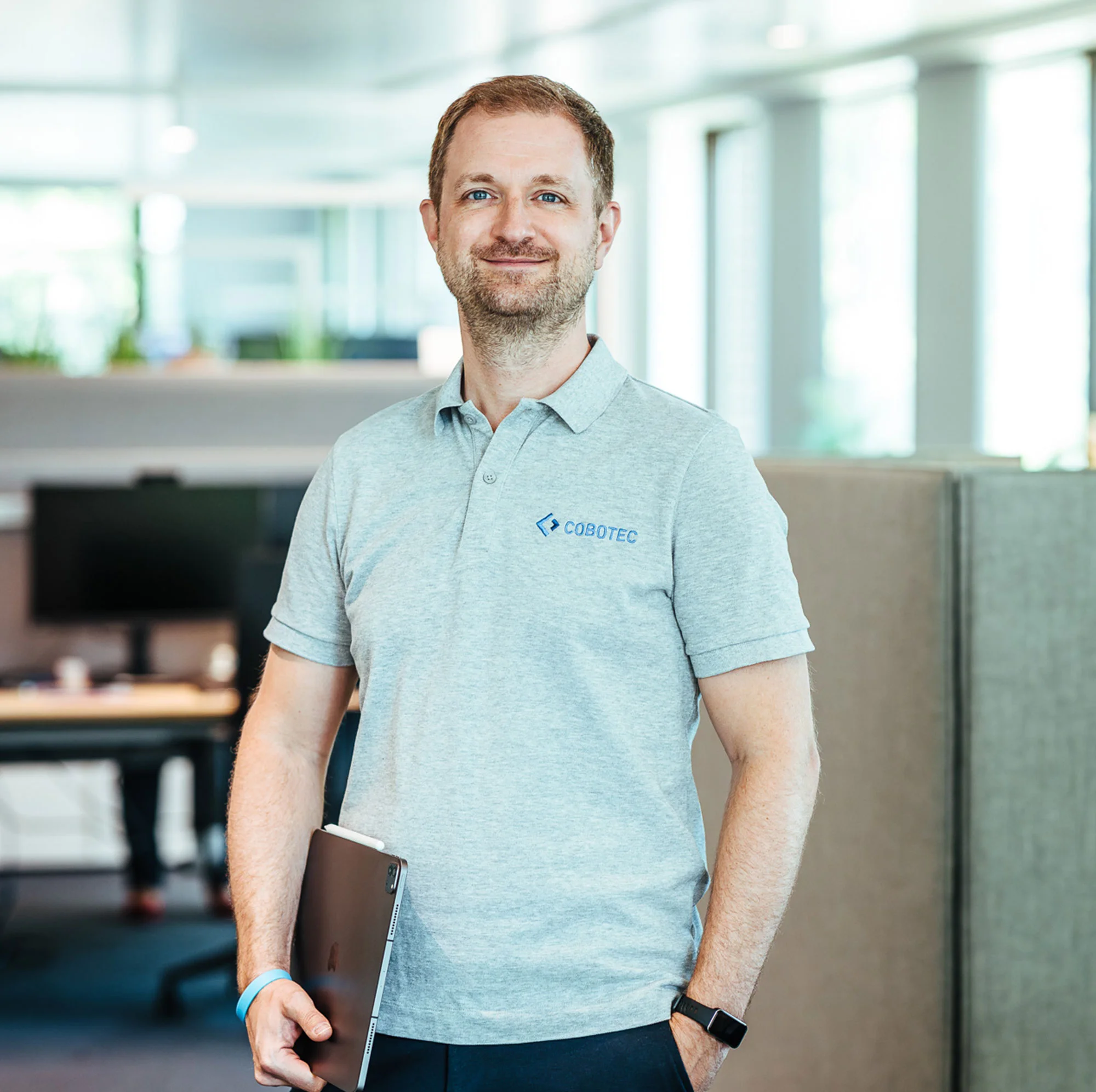 Mann steht in einem modernen Büro und hält ein Tablet in der Hand, trägt ein graues Polo-Shirt mit dem Logo „COBOTEC“.